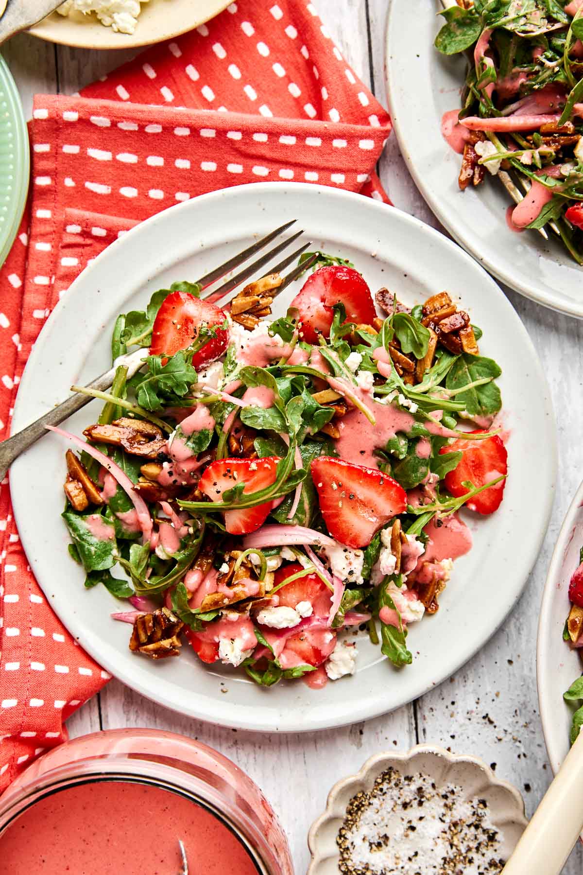 A white plate with a fresh salad of arugula, sliced strawberries, goat cheese, candied almonds, and red onions, drizzled with pink dressing. A fork rests on the plate, and a red napkin is beside it.