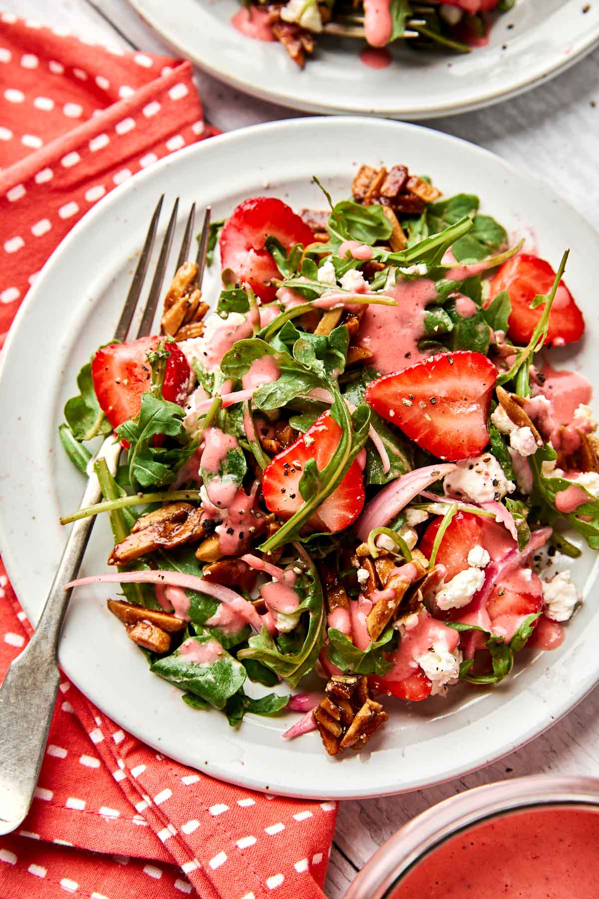 A fresh salad with sliced strawberries, arugula, almonds, red onions, and crumbled cheese, drizzled with pink dressing, served on a white plate with a fork on a red polka dot napkin.