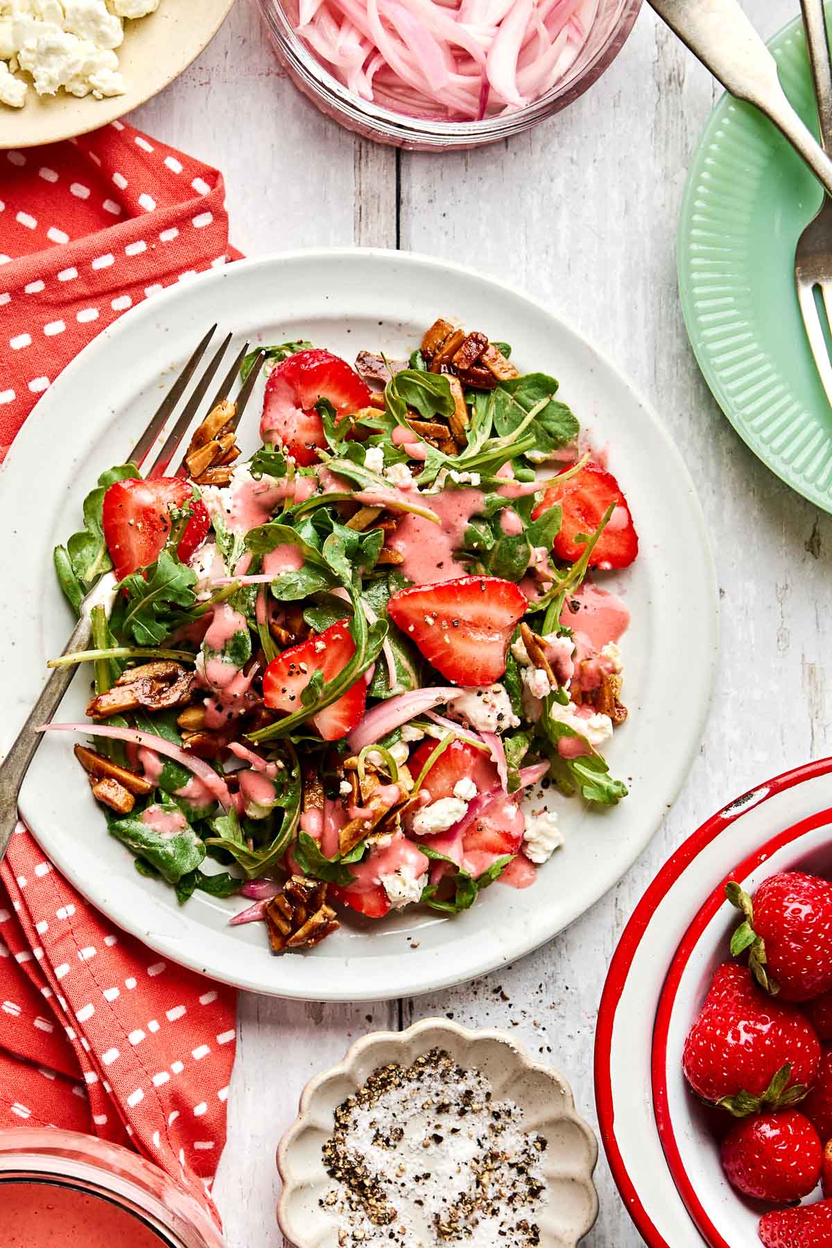 A plate of fresh salad with arugula, sliced strawberries, almonds, and crumbled cheese, drizzled with pink dressing, sits on a white table beside a fork, a red napkin, and bowls of strawberries and pink pickled onions.