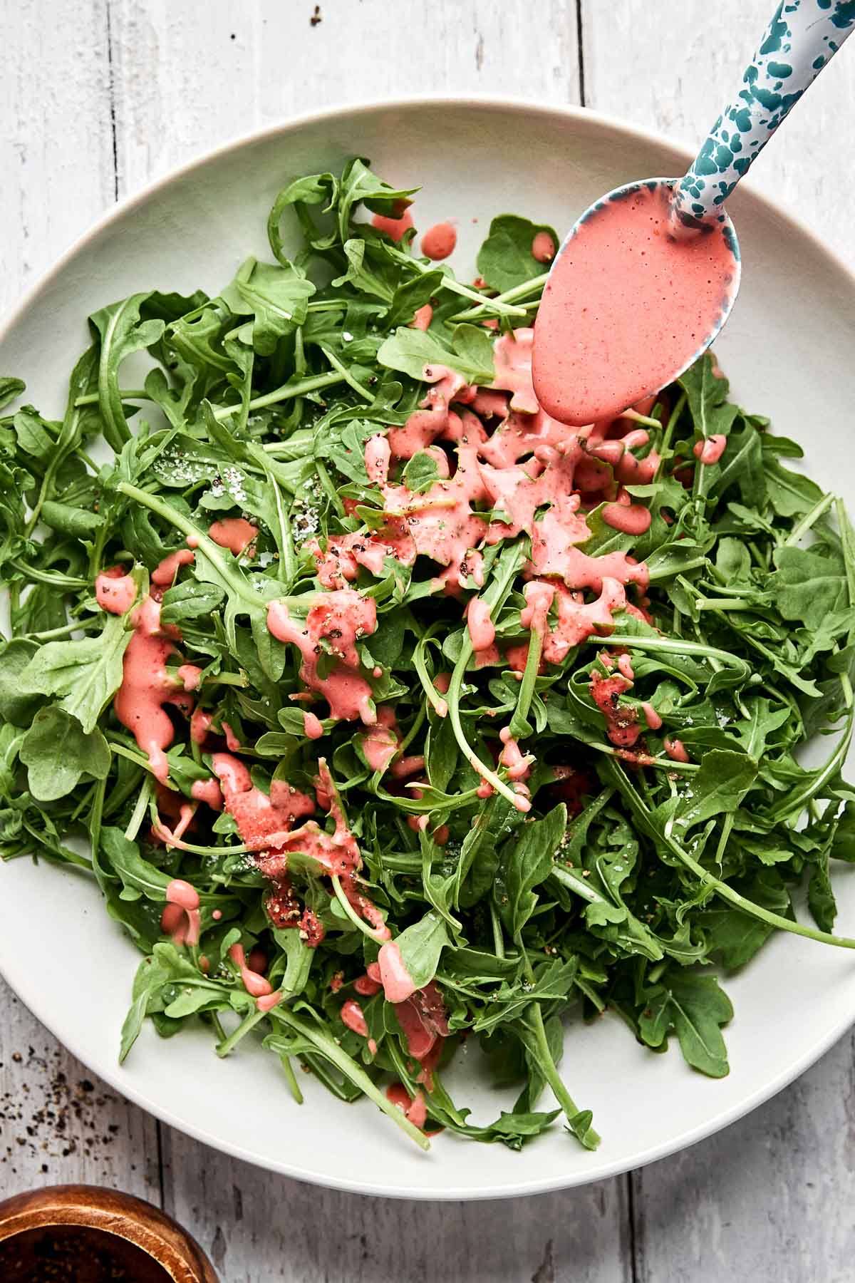 A bowl of fresh arugula leaves being drizzled with a creamy pink salad dressing from a spoon above.