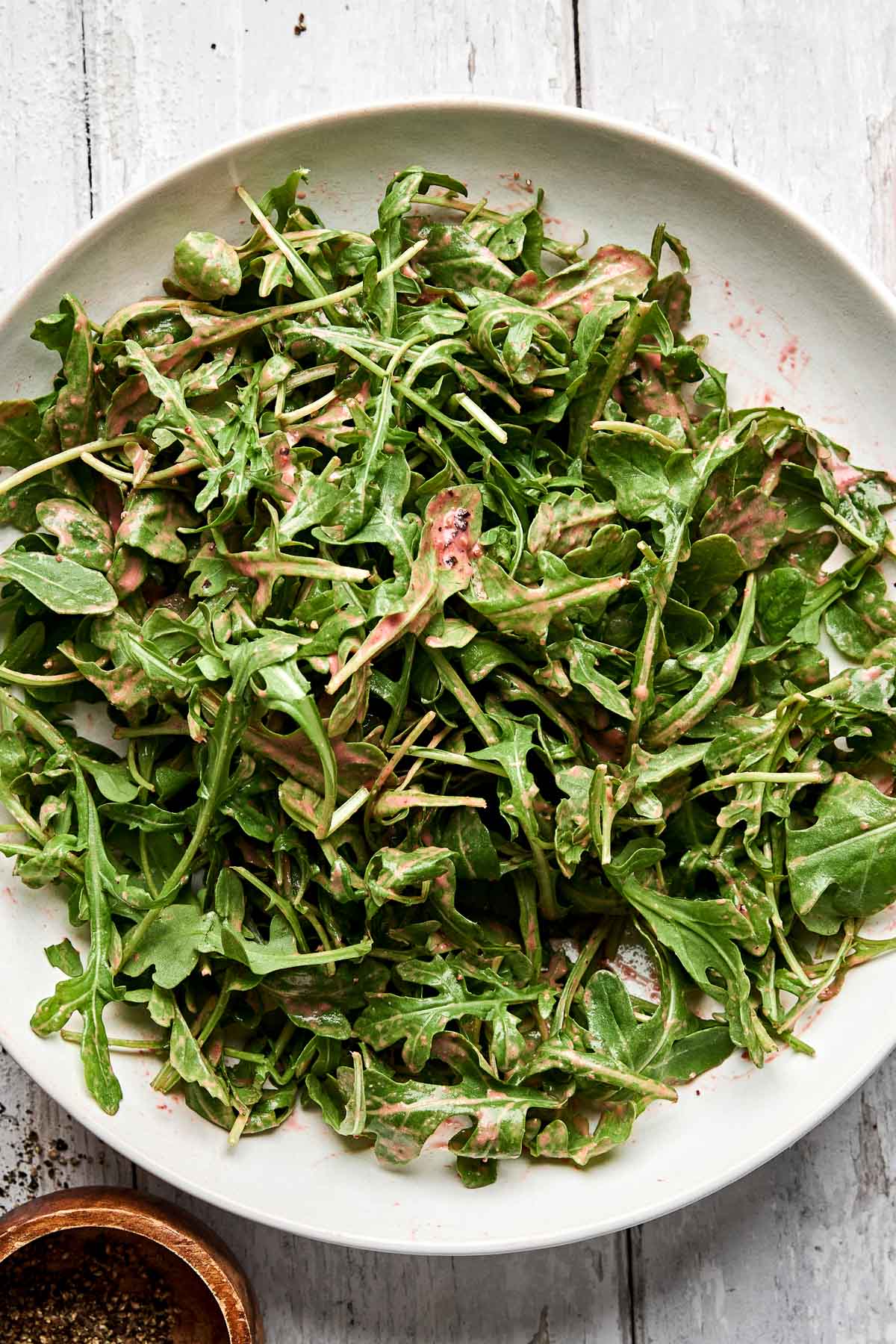 A plate of fresh arugula salad tossed with a creamy pink dressing sits on a rustic white wooden table. A small bowl of ground black pepper is visible in the corner.