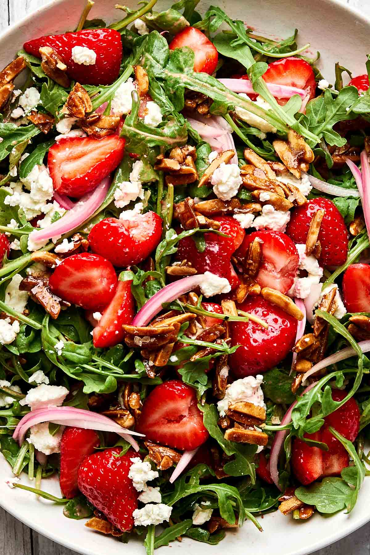 A close-up of a fresh salad with arugula, sliced strawberries, crumbled goat cheese, red onion, and candied almonds on a white plate.