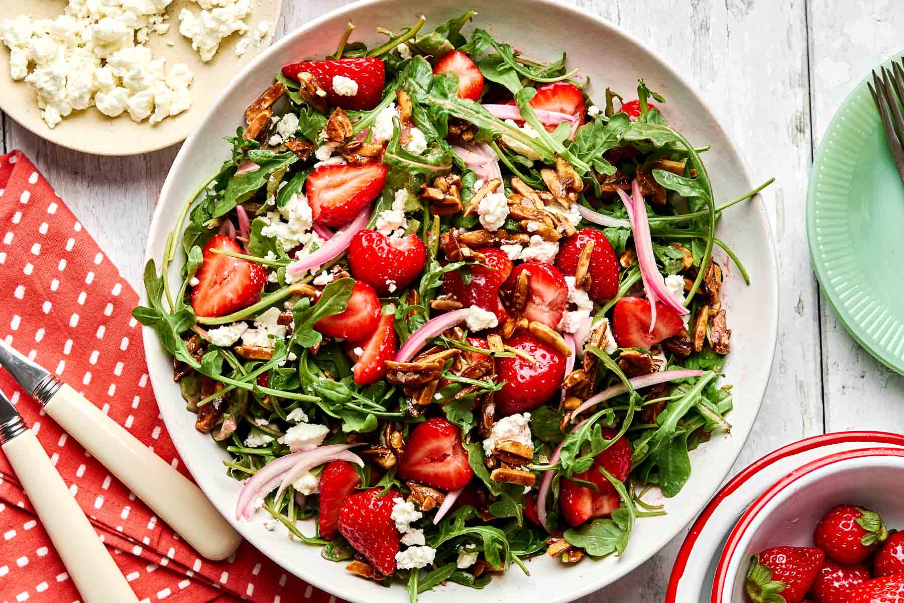 A bowl of arugula salad with sliced strawberries, red onions, crumbled feta cheese, and candied pecans, set on a white table with a red napkin, utensils, and small plates nearby.