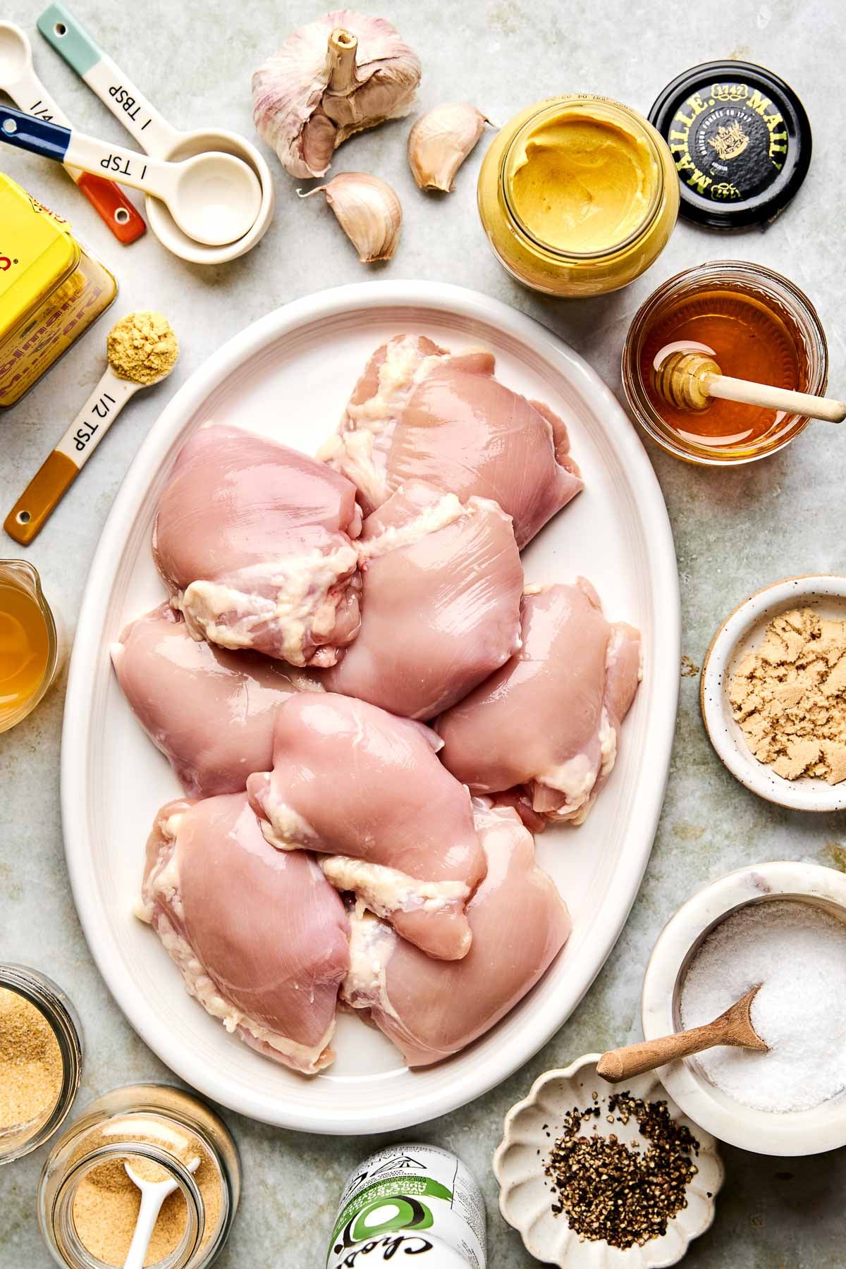 Raw chicken thighs on a white platter surrounded by ingredients in bowls and jars, including garlic, mustard, honey, brown sugar, spices, olive oil, and measuring spoons, arranged neatly on a light countertop.