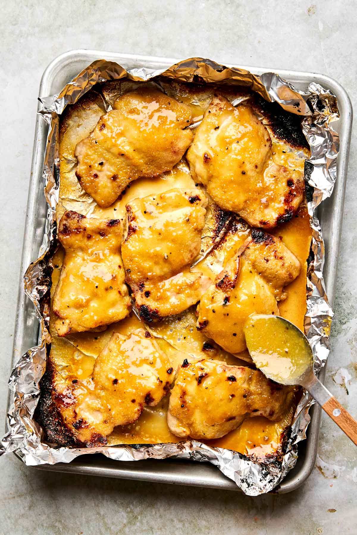 Baked chicken thighs covered in a golden, glossy sauce on a foil-lined baking sheet, with a spoon drizzling sauce over one piece.
