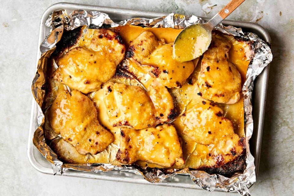 Baked chicken thighs covered in a golden, creamy sauce on a foil-lined baking sheet. A spoon is drizzling more sauce over the chicken.