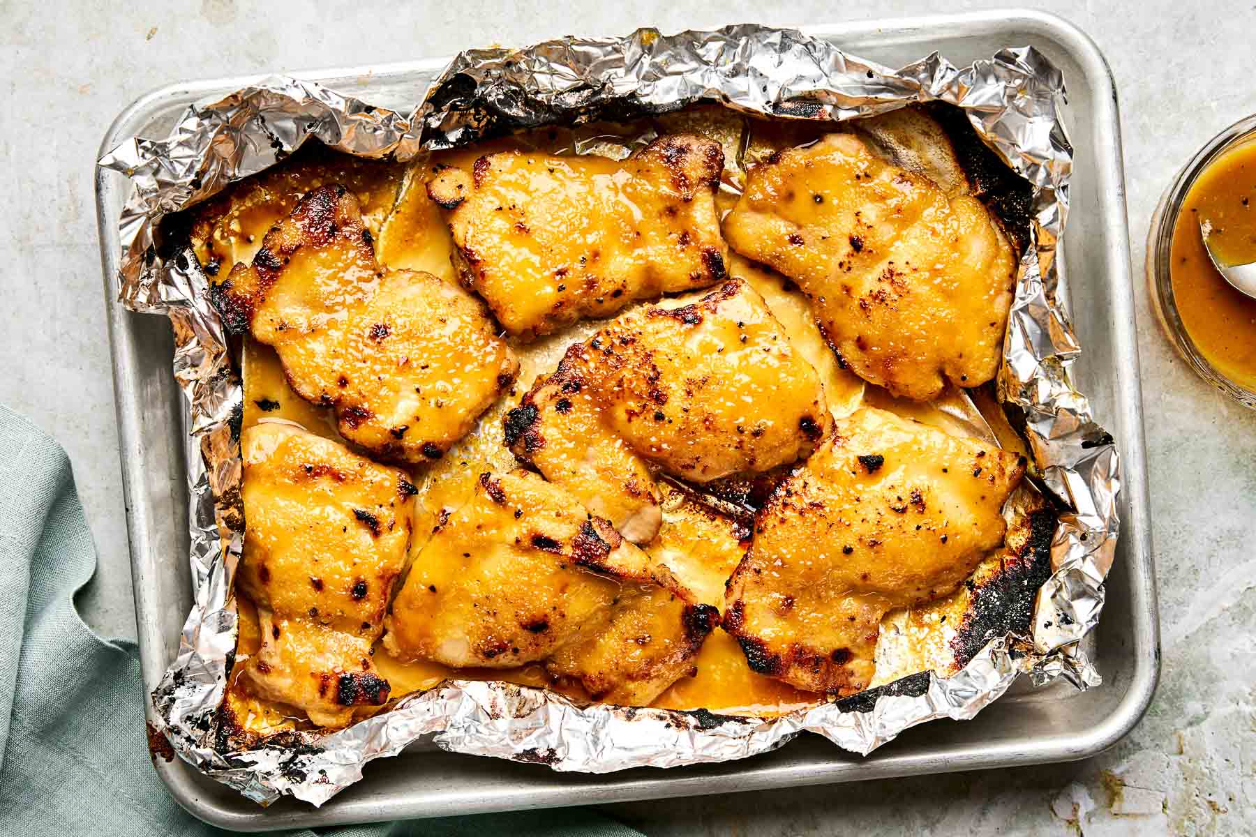Baked chicken thighs coated in a golden, slightly crispy glaze, arranged on a foil-lined baking sheet, with a small dish of sauce nearby.