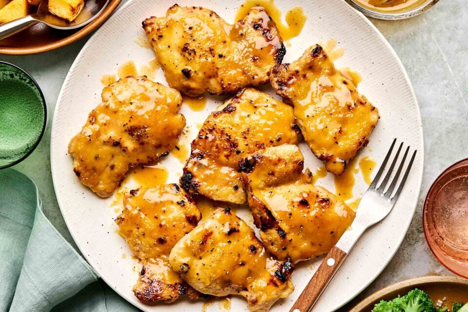 Seven pieces of grilled chicken coated in golden honey mustard sauce on a white plate, with a fork beside them and side dishes partially visible around the edge.