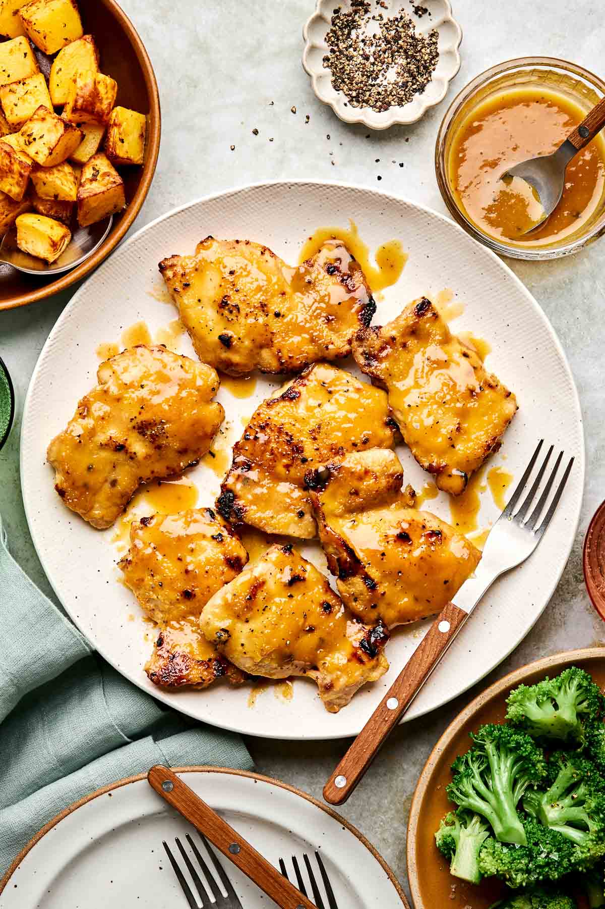 Grilled chicken thighs with a golden sauce on a plate, surrounded by a fork, roasted potatoes, steamed broccoli, black pepper, and a jar of extra sauce.