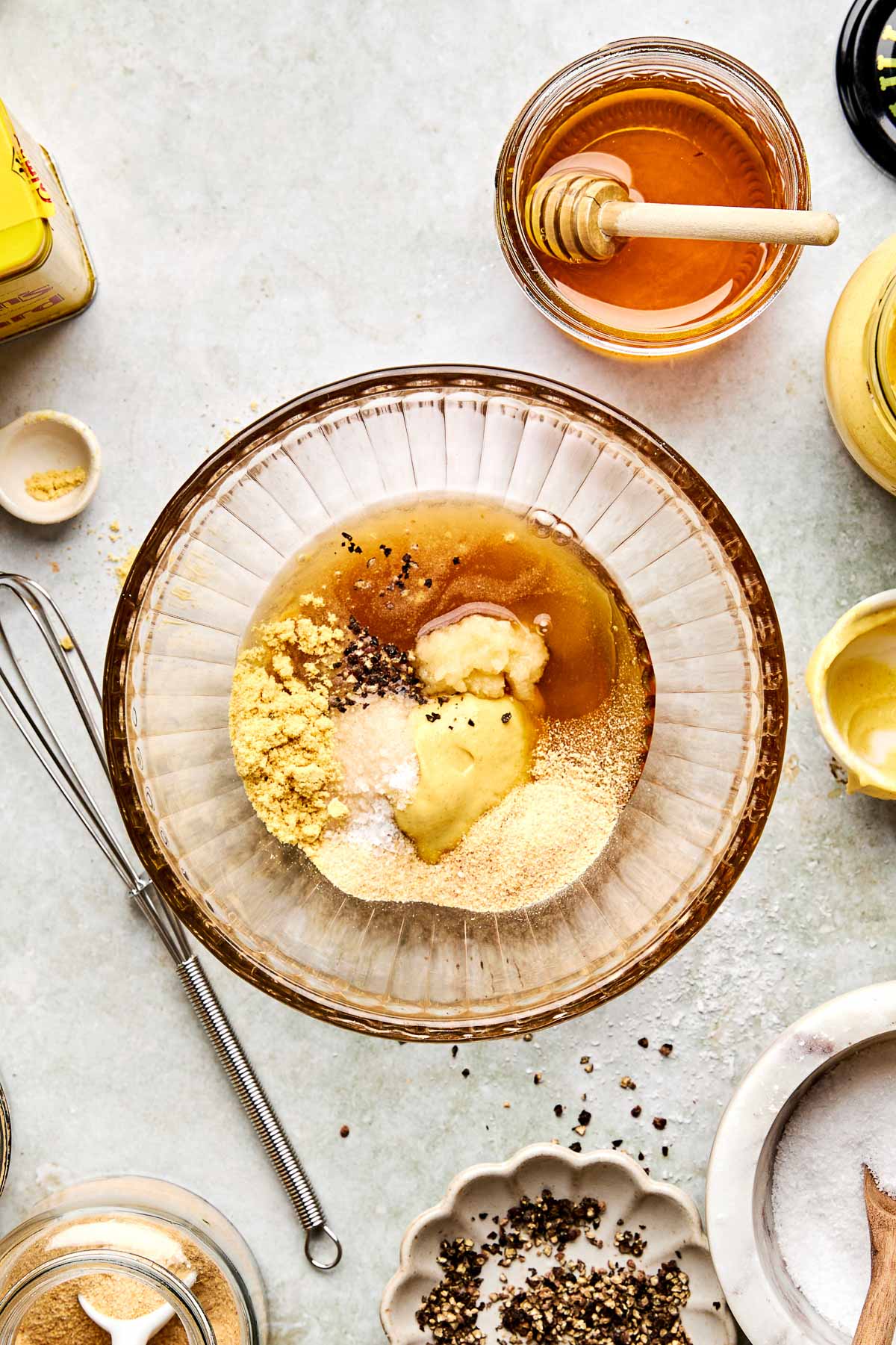 A glass bowl with mustard, honey, spices, and seasonings being mixed. Surrounding the bowl are a whisk, a small dish of cracked pepper, a jar of honey with a dipper, and jars of spices on a light countertop.