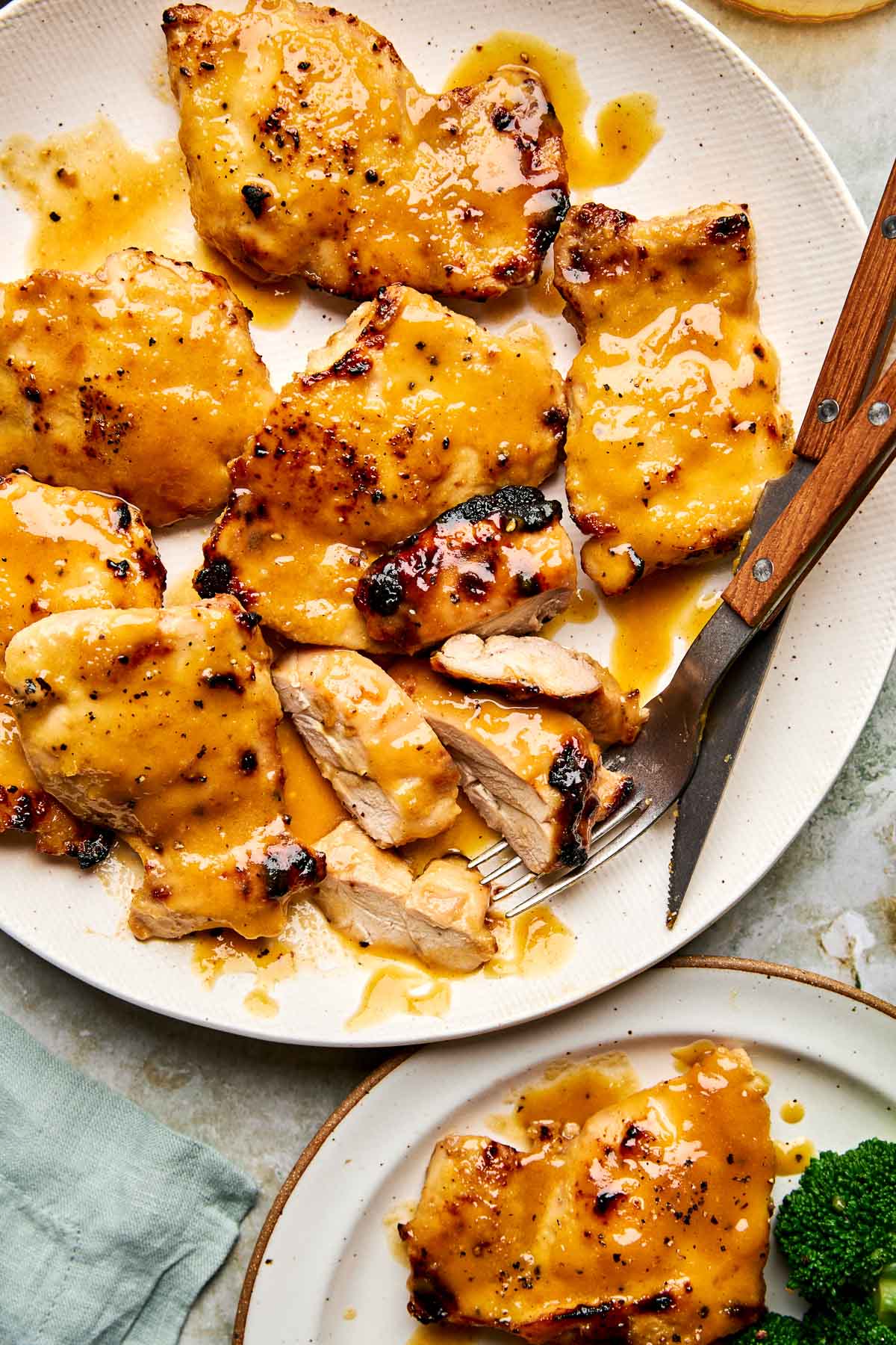 Plate of grilled chicken thighs covered in a golden honey mustard sauce, with a fork and knife beside a cut piece. A smaller plate with chicken and broccoli is partially visible.