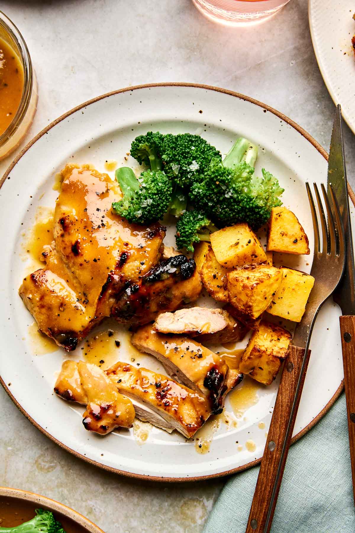 A plate with glazed chicken, roasted potatoes, and steamed broccoli. A fork and knife rest on the plate, and a light green napkin is nearby. The food is garnished with black pepper.