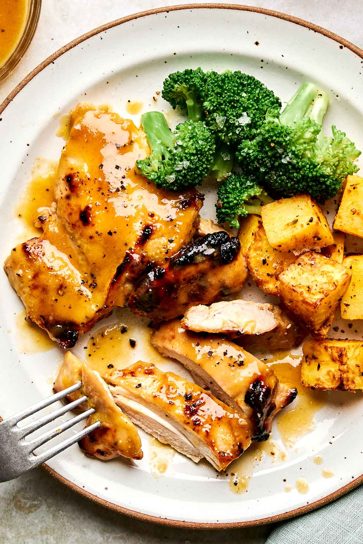 A plate with glazed grilled chicken, roasted potato cubes, steamed broccoli, and a fork, all drizzled with a golden sauce.