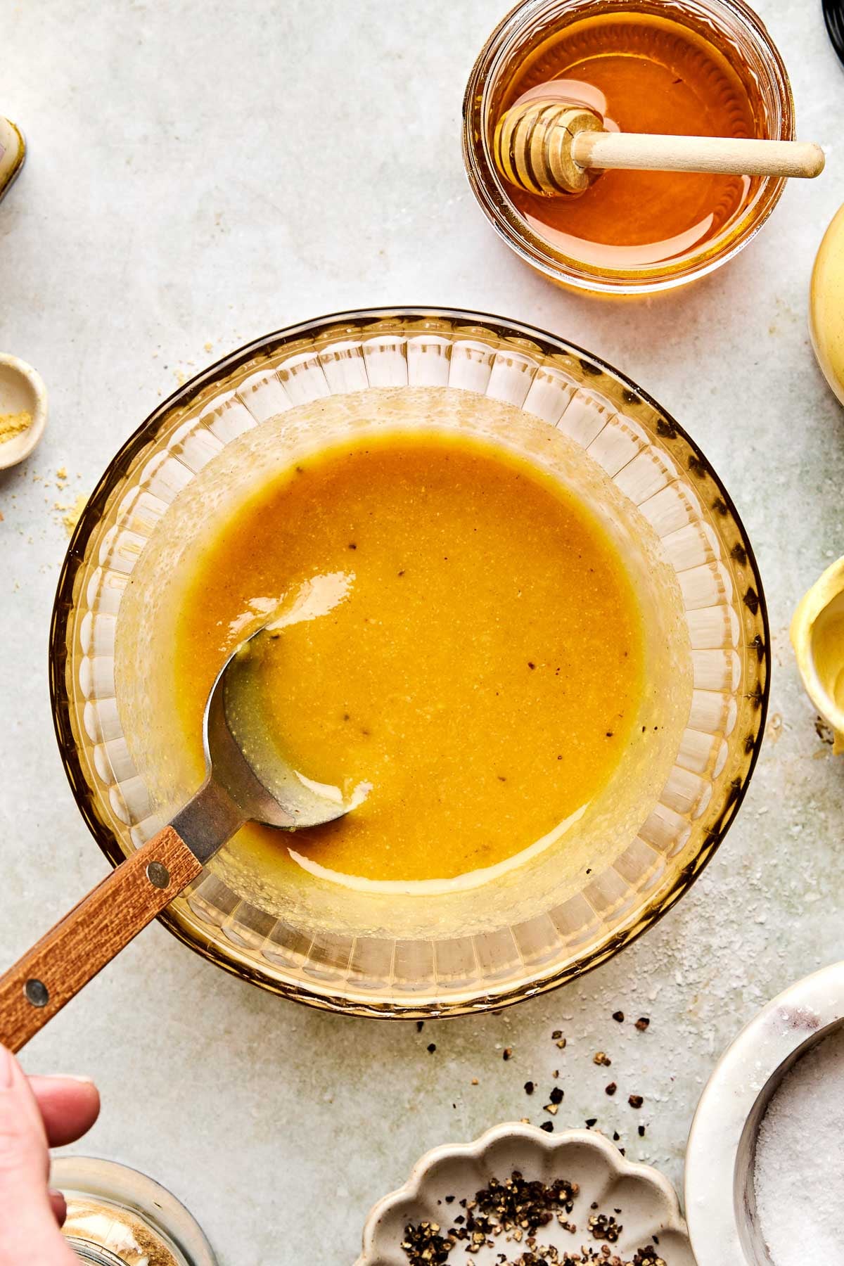 A hand holds a spoon mixing yellow salad dressing in a glass bowl, surrounded by ingredients like honey, lemon, and pepper on a light countertop.
