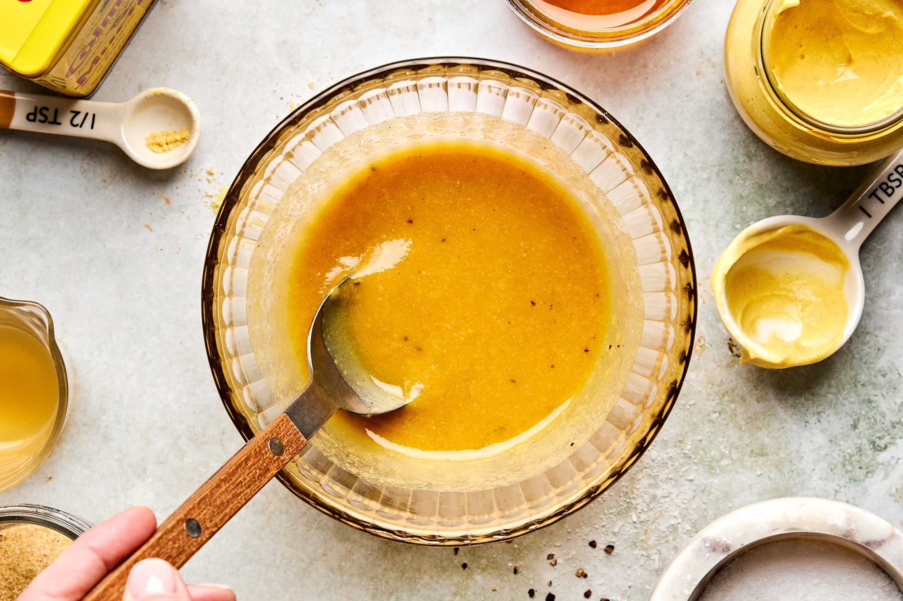 A hand stirs yellow salad dressing in a glass bowl with a spoon, surrounded by measuring spoons, mustard, honey, and seasonings on a light countertop.