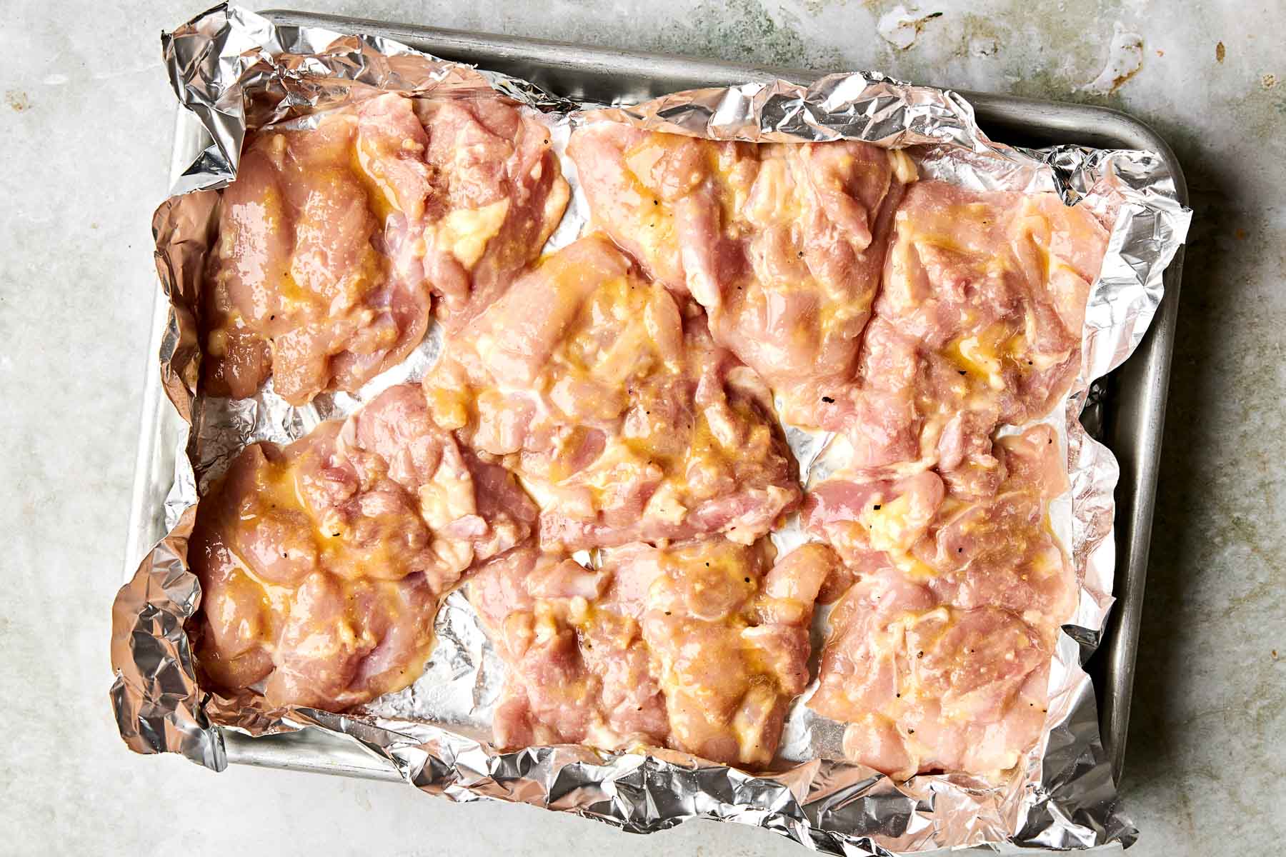 Raw, marinated chicken pieces are spread out on a foil-lined baking sheet, ready to be cooked.