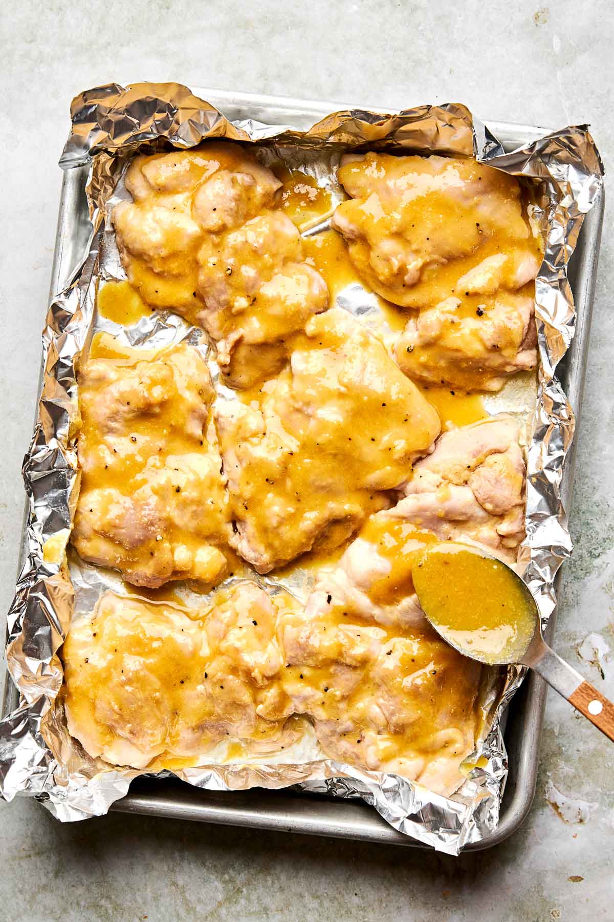 Baked chicken thighs covered in a golden sauce on a foil-lined baking sheet. A spoon with sauce rests on the edge of the pan.