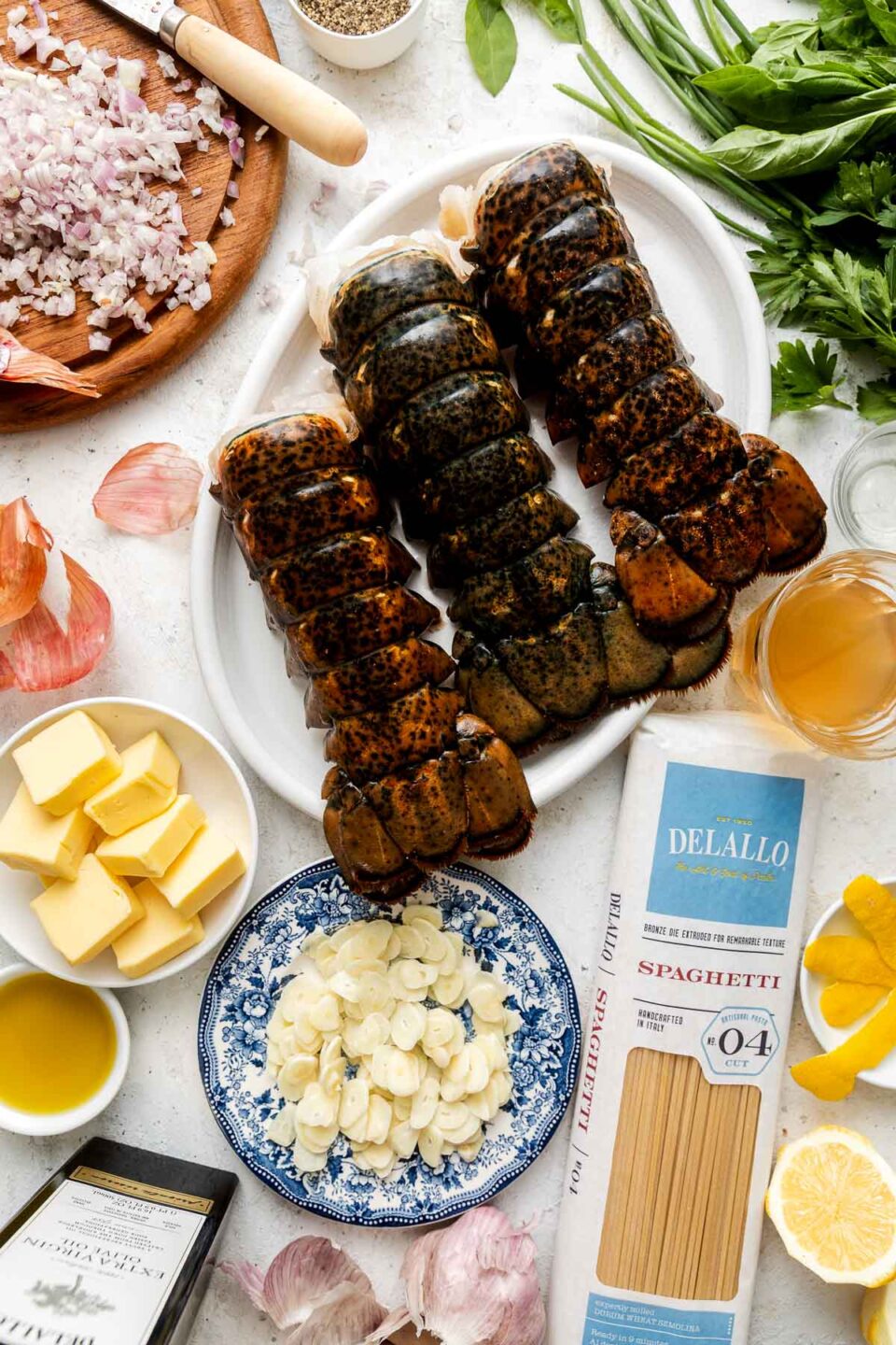 Three raw lobster tails on a plate, surrounded by chopped shallots, garlic, butter, lemon, spaghetti, herbs, oil, and broth, displayed on a white surface ready for cooking.