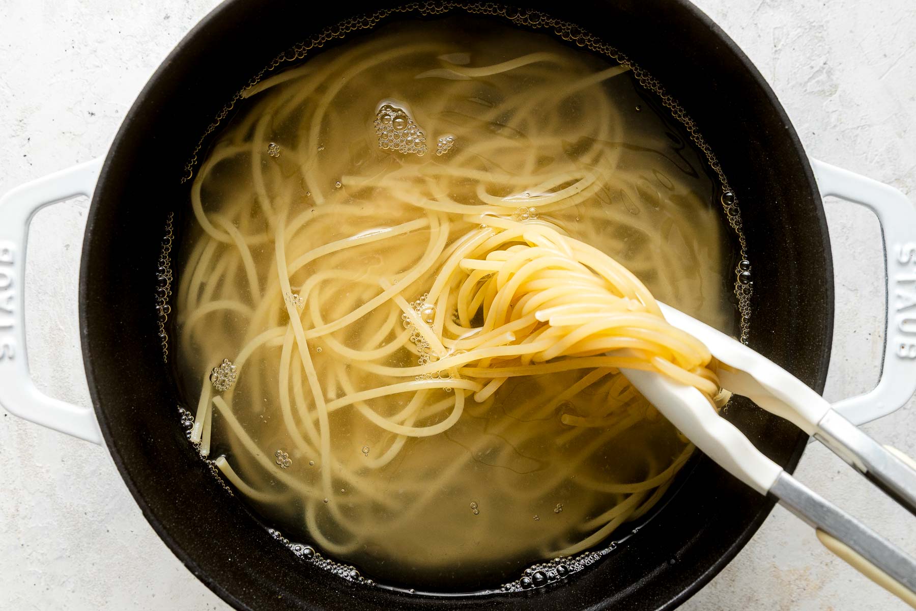 A pot of boiling water with spaghetti noodles being lifted out using metal tongs.