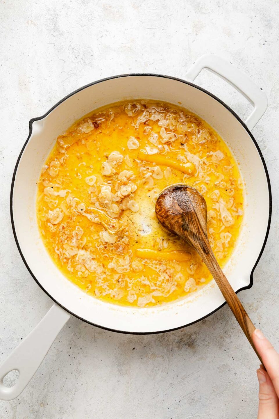 A hand stirs sliced garlic sautéing in melted butter in a white enameled pan on a light countertop.