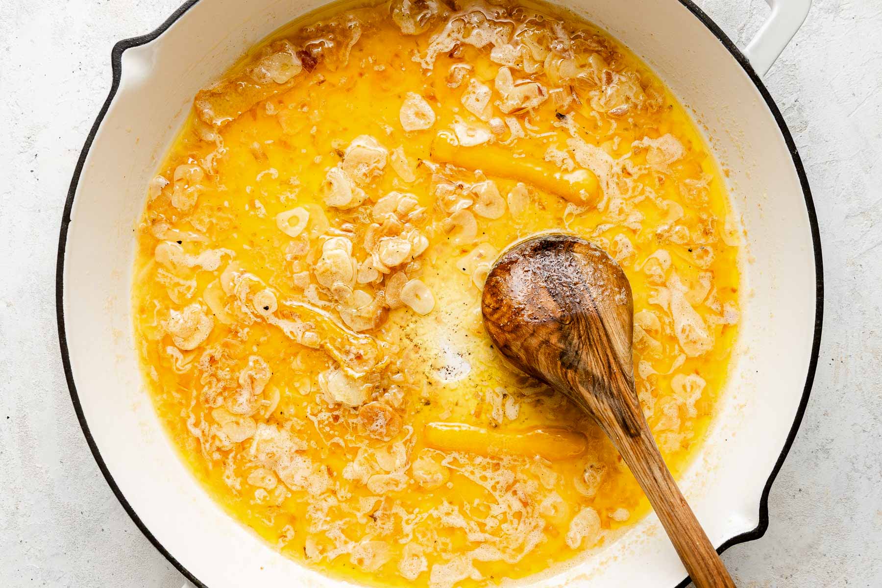 Sliced garlic is sautéed in melted butter in a white pan. A wooden spoon rests inside, stirring the golden mixture.