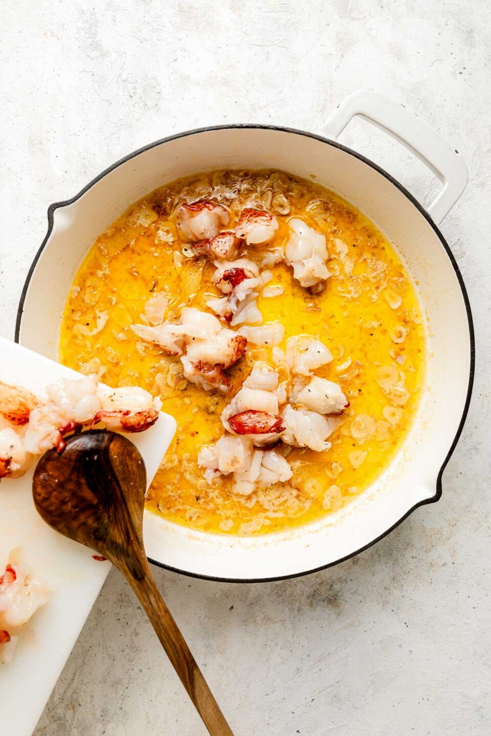 Chopped lobster tails being added from a white cutting board into a pot with melted butter, garlic, and shallots, using a wooden spoon.