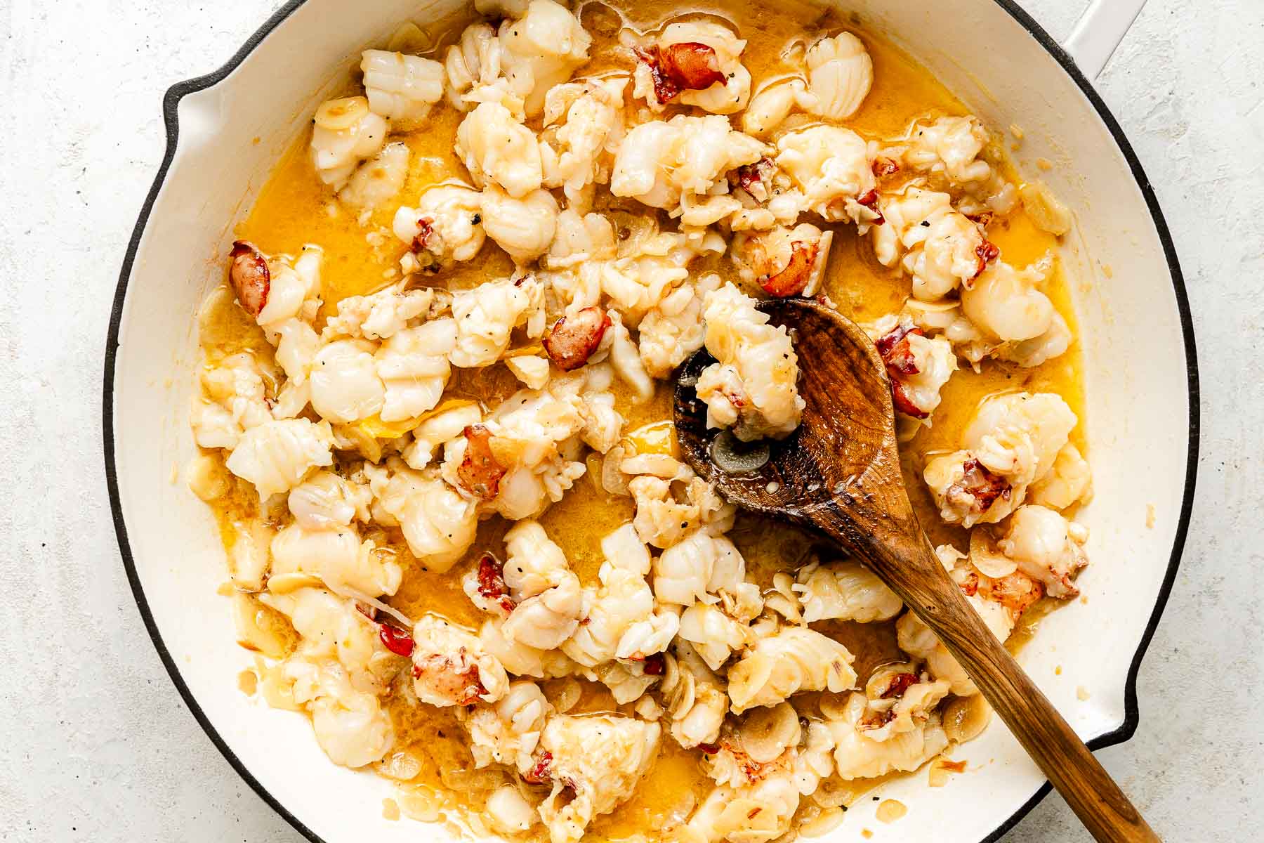 Chopped lobster pieces cooking in a creamy sauce in a white skillet, stirred with a wooden spoon.