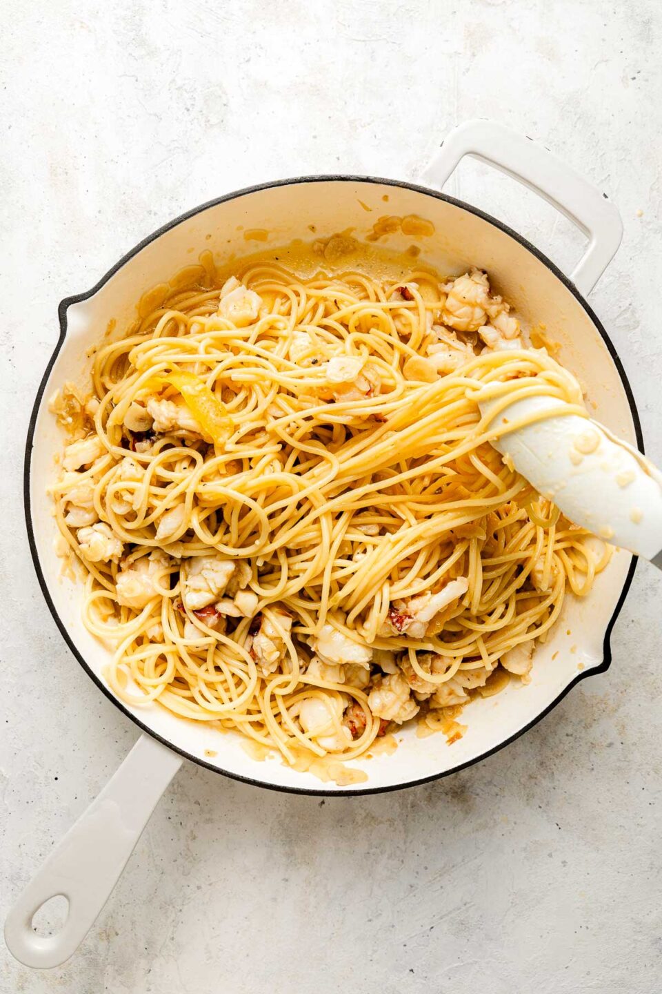 Spaghetti and chunks of lobster are mixed together in a white pan with a white handle, resting on a light-colored countertop. A white spatula is lifting some of the pasta.