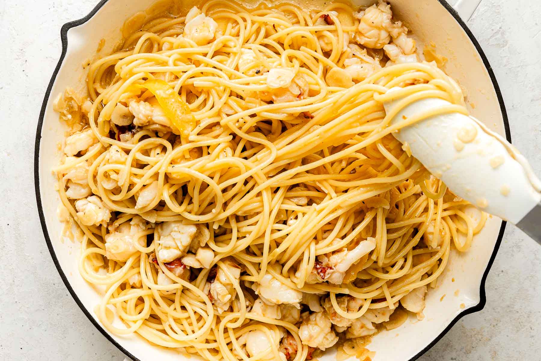 Cooked spaghetti tossed with chunks of lobster in a creamy sauce, served in a white skillet, with tongs lifting some of the pasta.