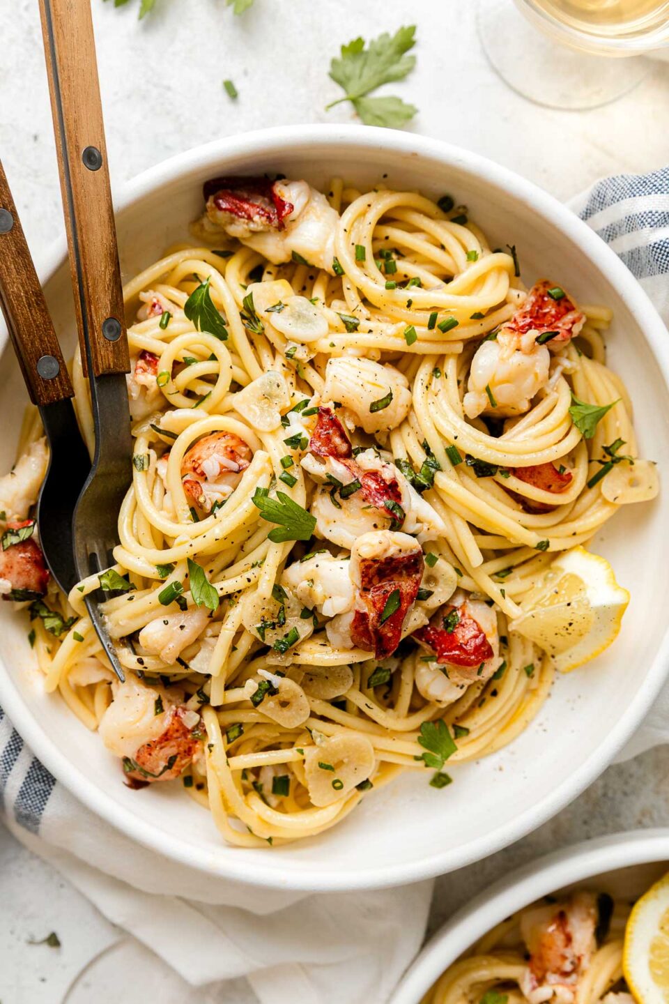 A bowl of spaghetti topped with lobster pieces, parsley, lemon slices, and a creamy sauce, with a fork and spoon resting on the side.