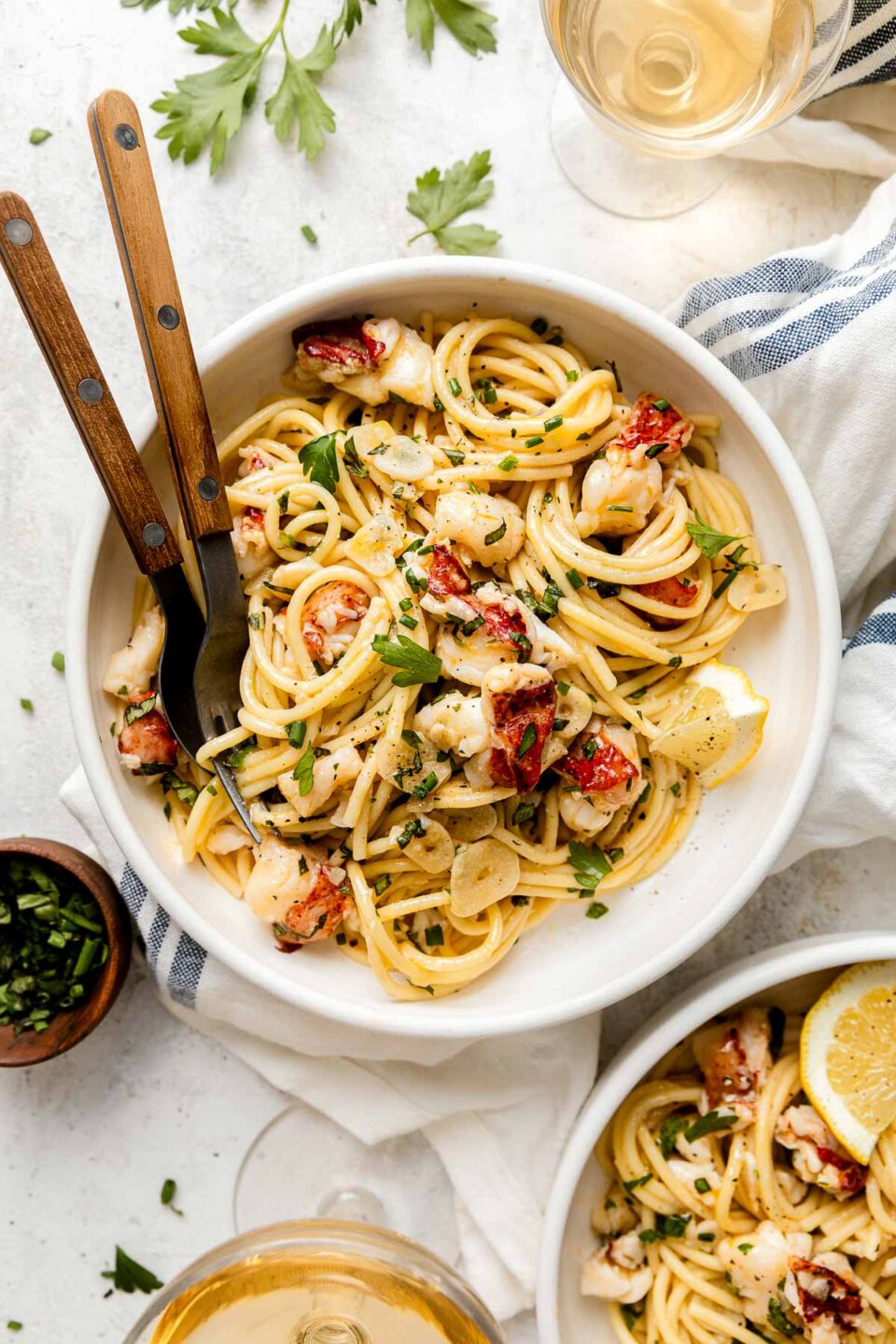 A bowl of pasta with lobster pieces, herbs, and lemon, served with two forks. Small bowls of herbs and lemon slices, a glass of white wine, and a striped napkin are nearby.