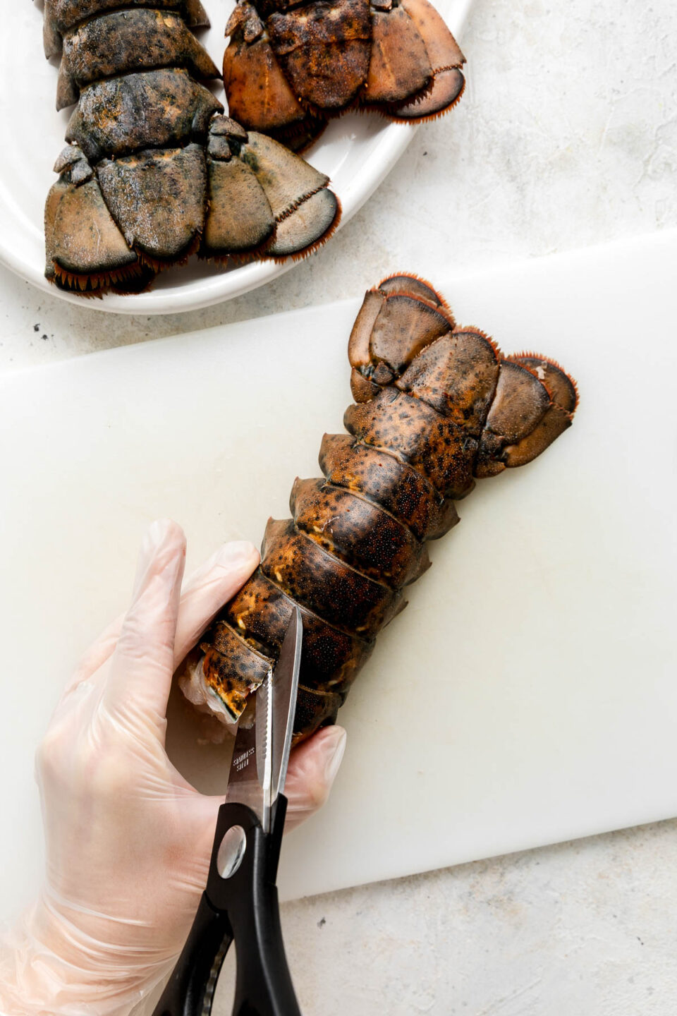 A gloved hand uses kitchen shears to cut through the top shell of a raw lobster tail on a white cutting board, with more lobster tails on a plate in the background.