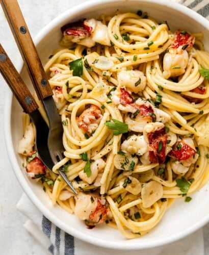 A bowl of pasta with lobster pieces, herbs, and garlic slices, served with a fork and knife on the side.
