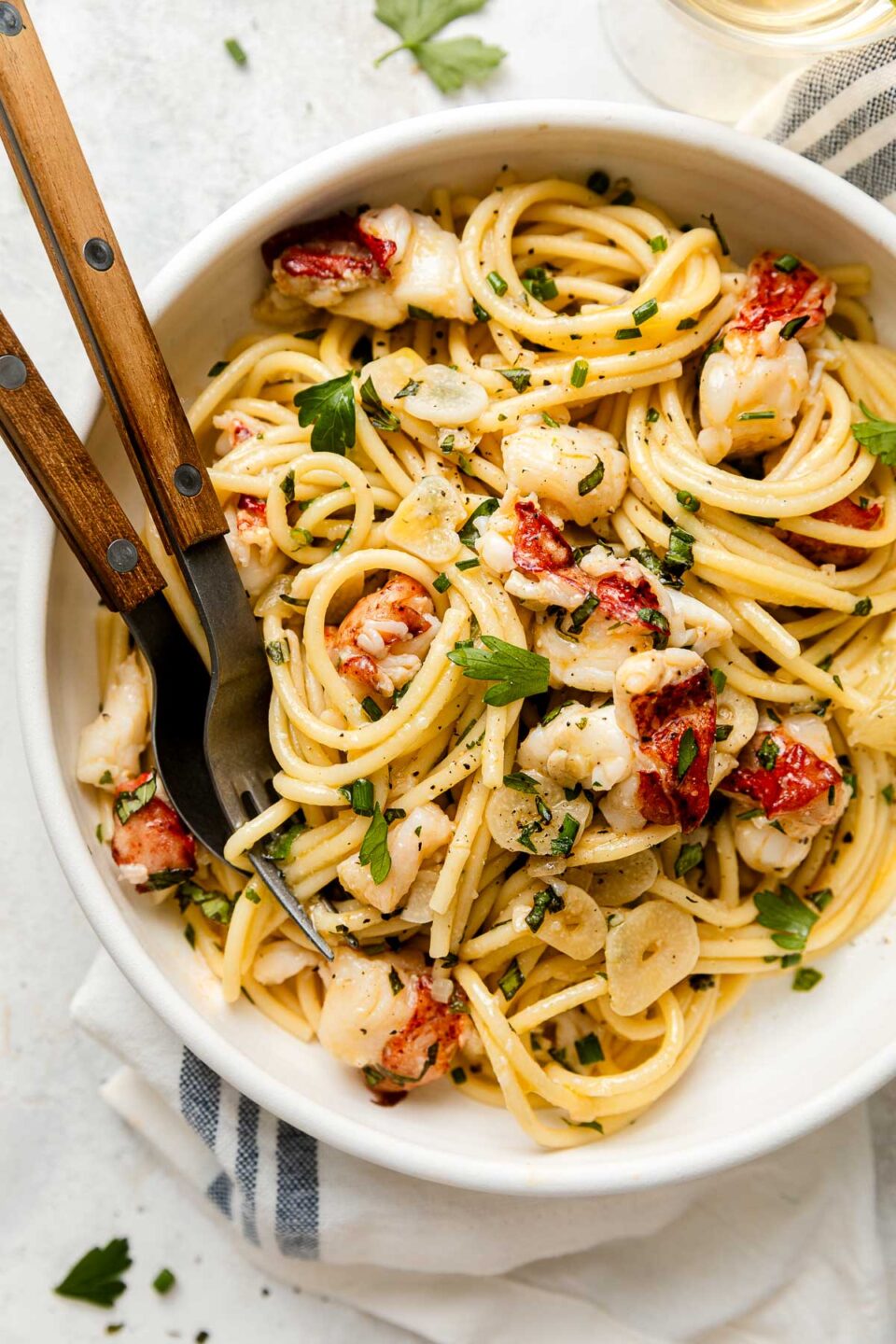 A bowl of pasta with lobster pieces, herbs, and garlic slices, served with a fork and knife on the side.