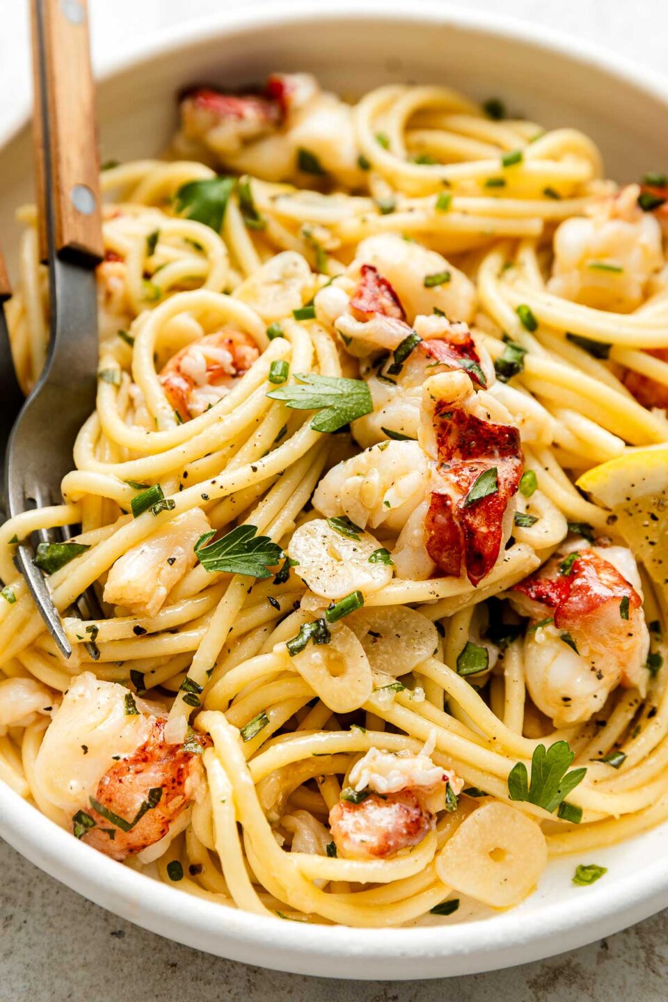 A bowl of pasta topped with lobster pieces, sliced garlic, fresh parsley, and black pepper, with a fork resting on the side.