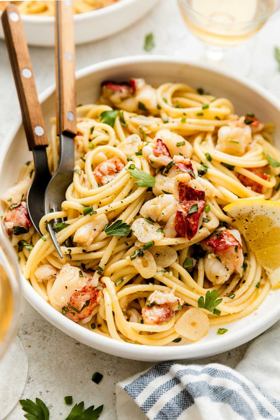A bowl of linguine pasta with pieces of lobster, slices of garlic, fresh herbs, and a lemon wedge. Two wooden-handled utensils rest in the bowl, and glasses of white wine are nearby on a light surface.