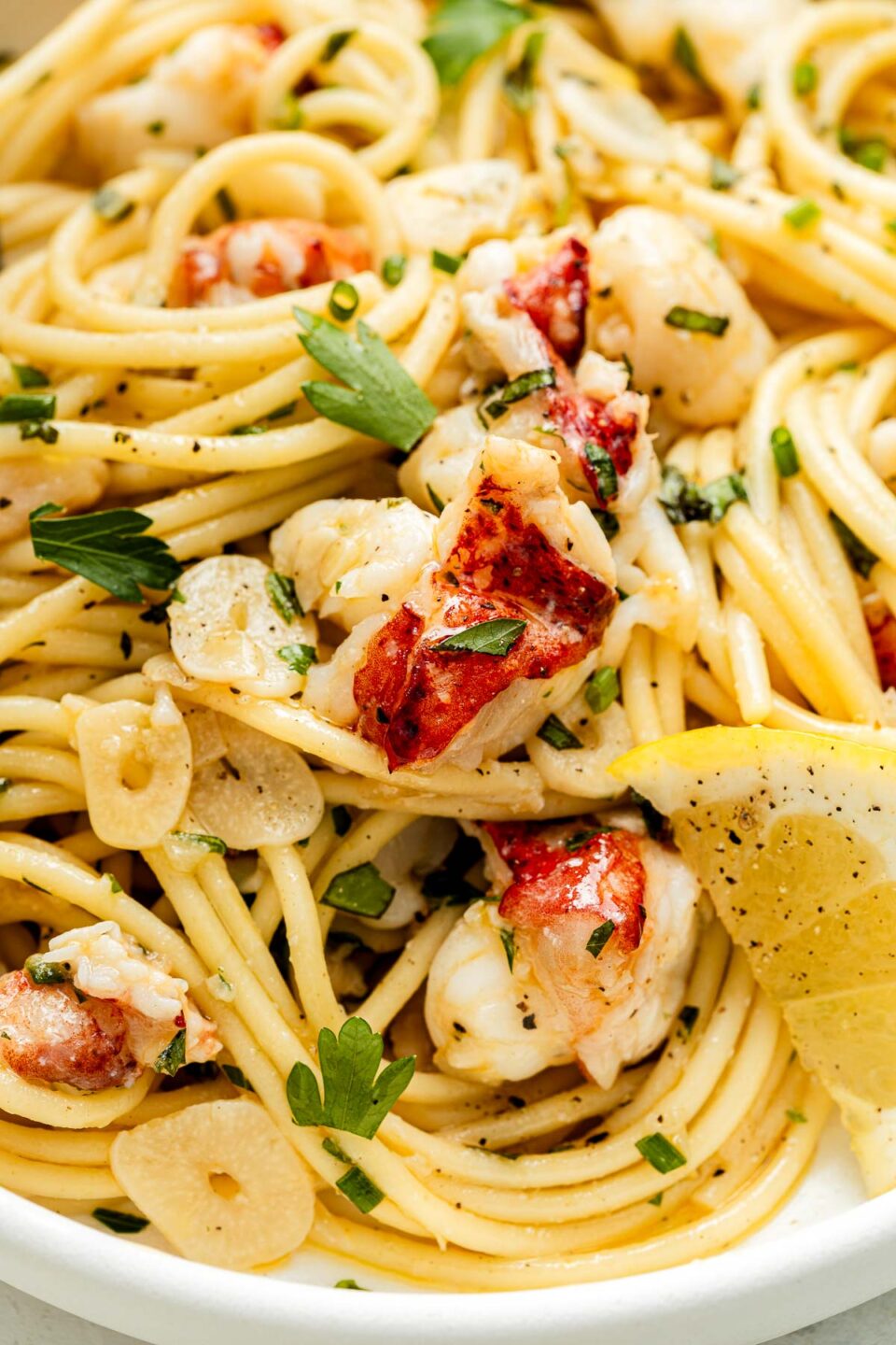 Close-up of spaghetti pasta with lobster chunks, sliced garlic, fresh parsley, lemon wedges, and black pepper, all tossed together in a light sauce.