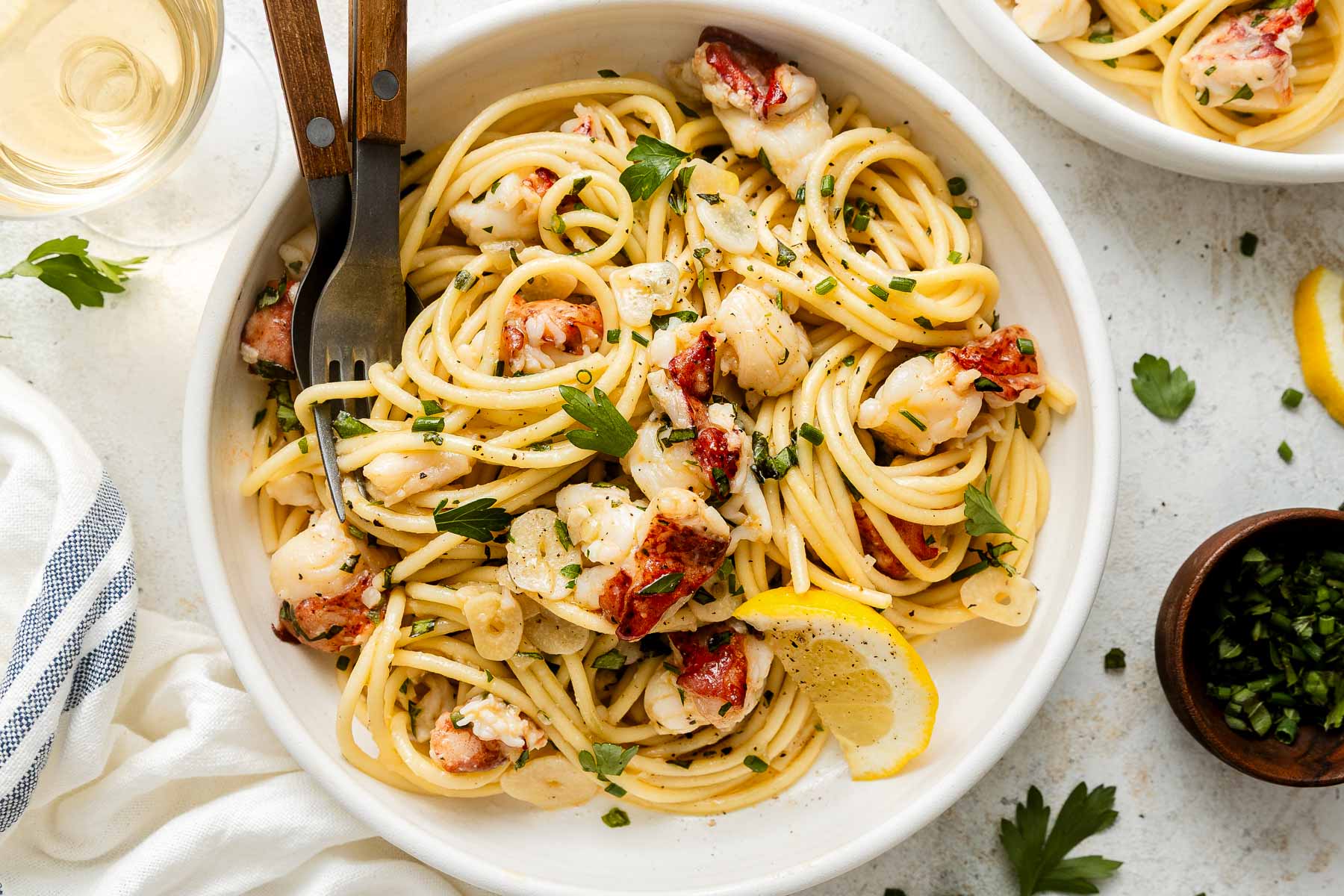 A bowl of pasta with lobster pieces, garlic, herbs, and a lemon wedge, served with a fork and garnished with parsley. A glass of white wine, napkin, and extra herbs are nearby.