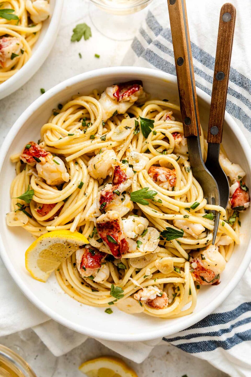 A bowl of pasta with chunks of lobster, garnished with parsley and lemon, with a fork and spoon resting inside. A striped napkin and another dish are partially visible beside the bowl.