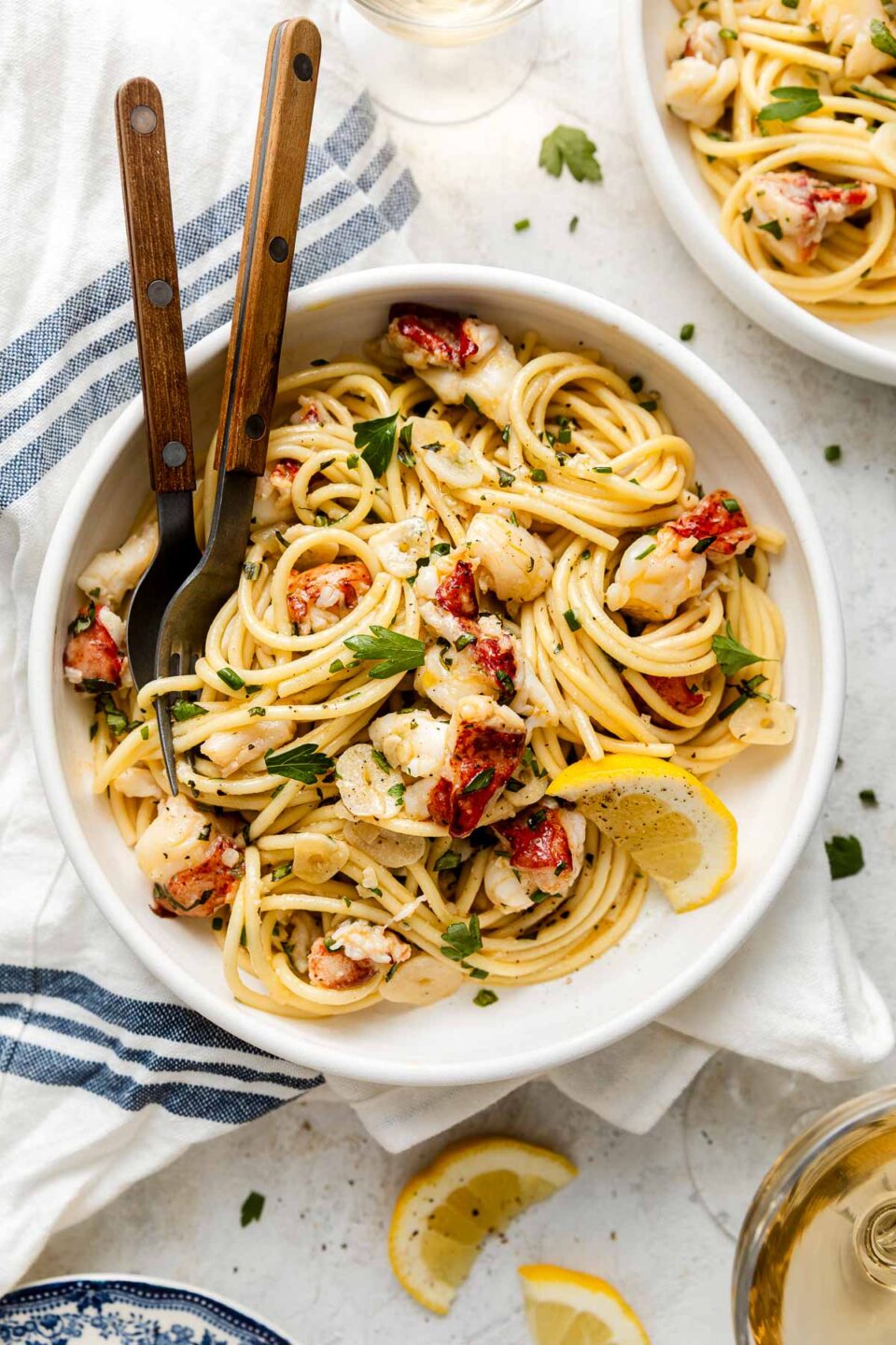 A bowl of spaghetti with lobster pieces, herbs, and lemon wedges, served with two wooden-handled utensils on a white tablecloth, next to a glass of white wine and a blue-striped napkin.