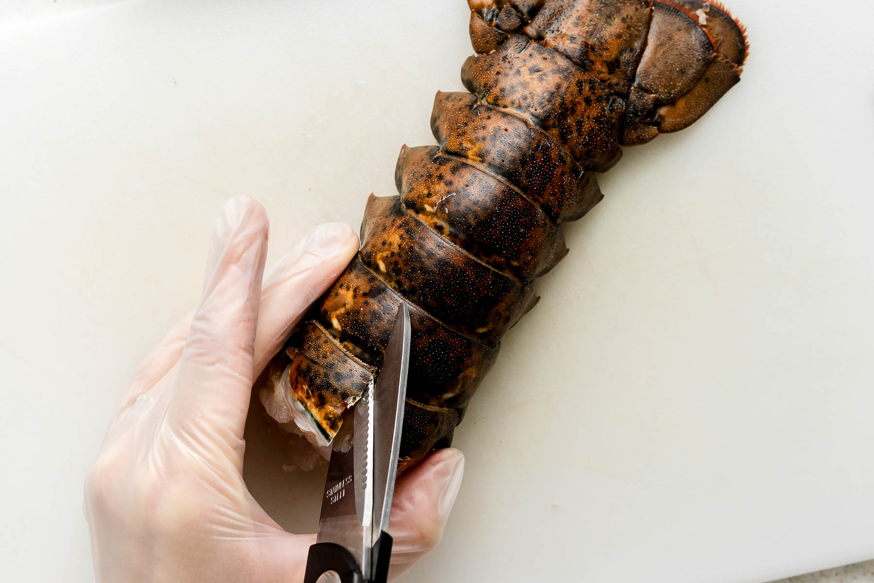 A gloved hand uses kitchen shears to cut through the shell of a raw lobster tail on a white cutting board.
