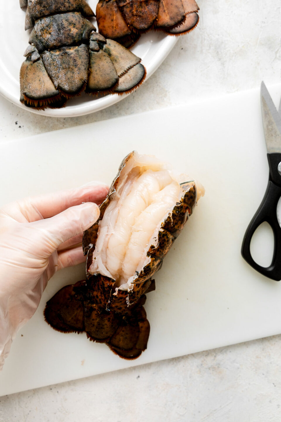 A gloved hand holds an opened raw lobster tail on a white cutting board, with kitchen scissors nearby and additional lobster tails on a plate in the background.