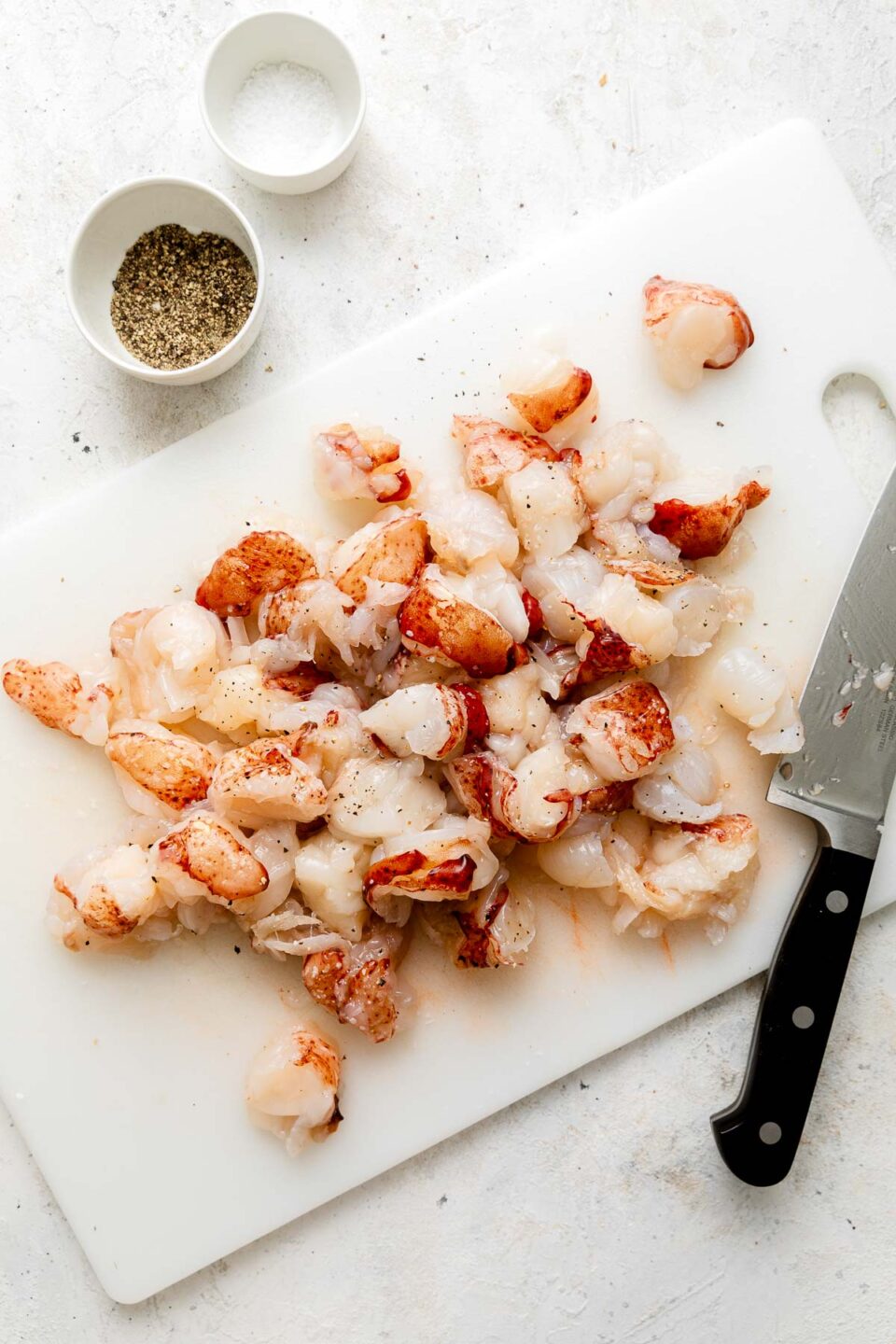 Chopped raw lobster meat is scattered on a white cutting board next to a black-handled knife. Two small bowls, one with black pepper and one with salt, are in the background.