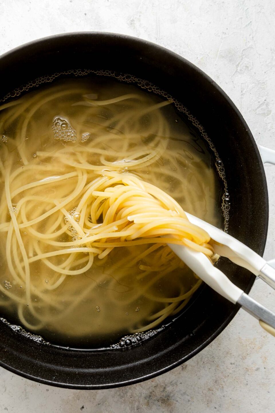 A pair of tongs lifts cooked spaghetti noodles from a pot filled with hot water.
