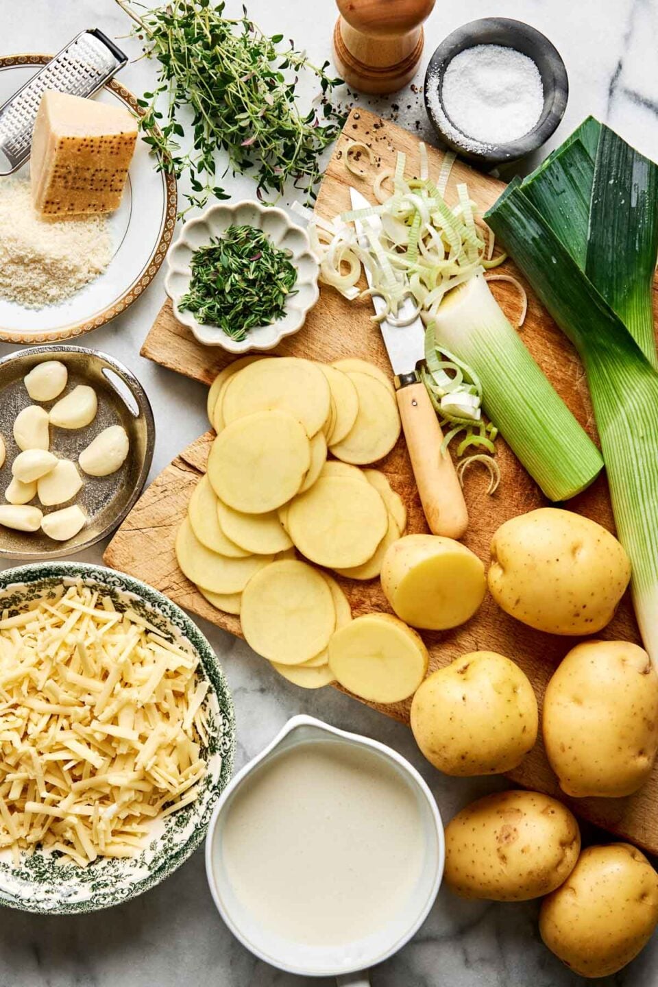 Ingredients for a potato leek dish including sliced potatoes, a whole leek, grated cheese, garlic cloves, thyme, a block of parmesan, salt, and a small pitcher of cream on a wooden cutting board.