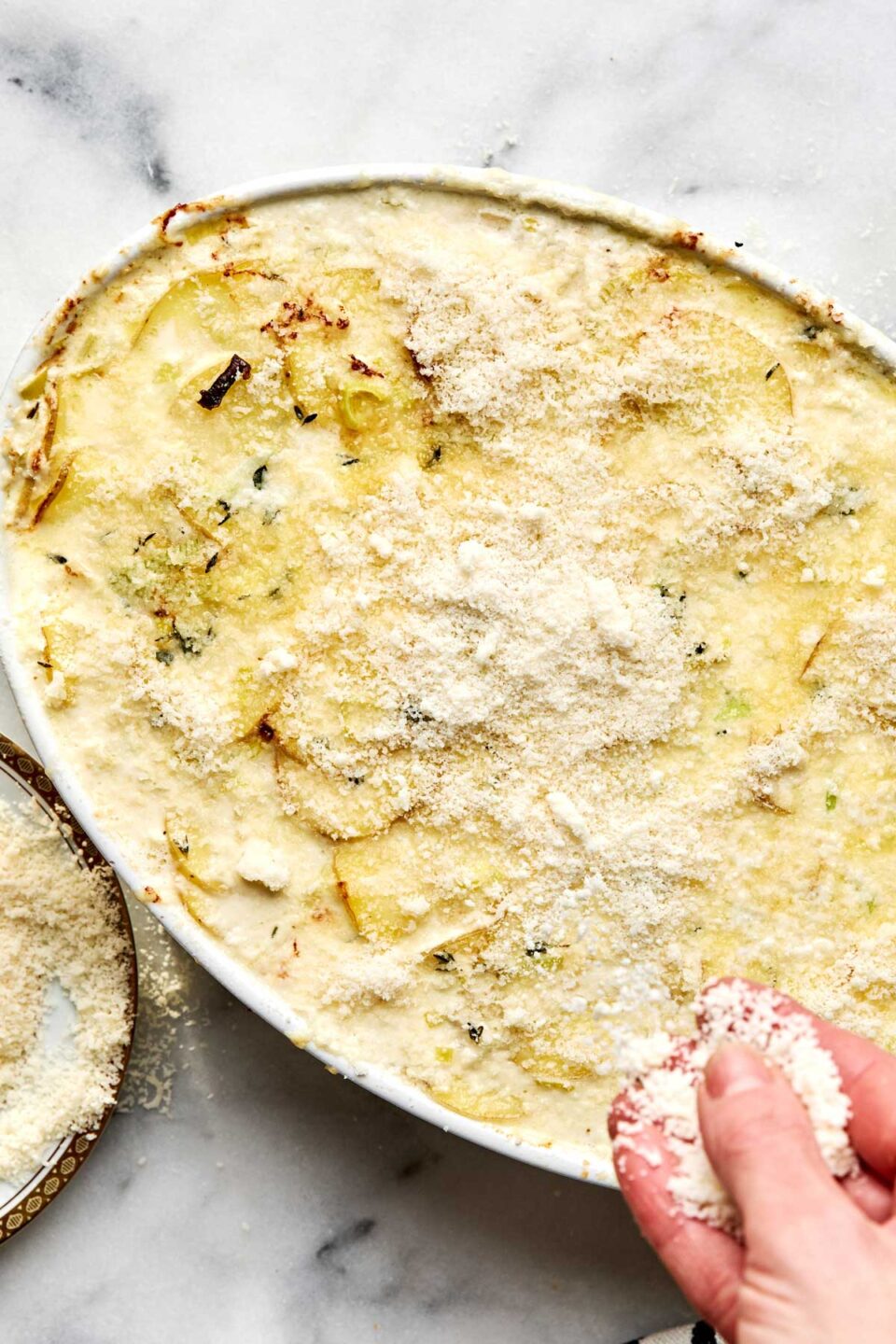 A hand sprinkles grated cheese over a casserole dish filled with au gratin potatoes.
