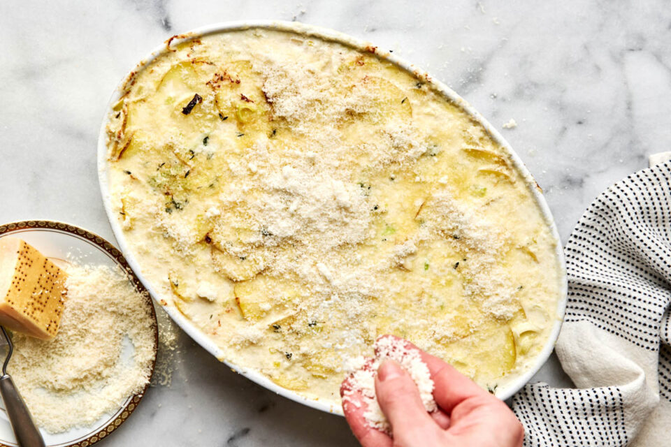 A hand sprinkles grated cheese over a baked potato gratin in an oval casserole dish.
