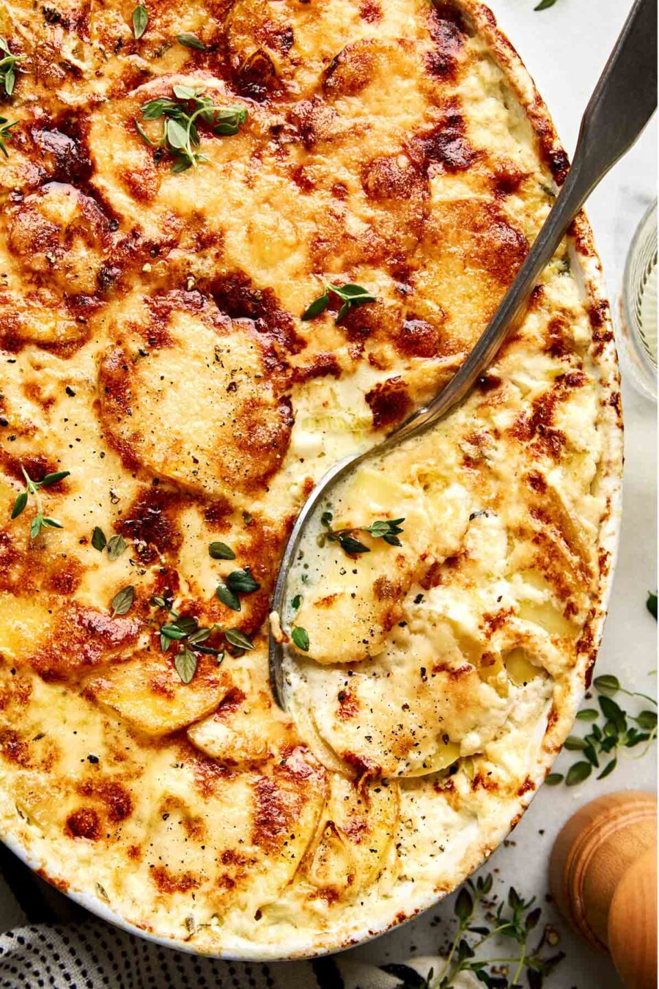 A serving spoon lifts a portion from a casserole dish filled with potatoes au gratin baked with a browned, cheesy crust and topped with fresh herbs.