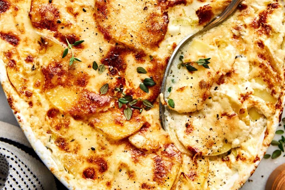 Baked potato gratin in a casserole dish, topped with fresh herbs and being scooped with a serving spoon.