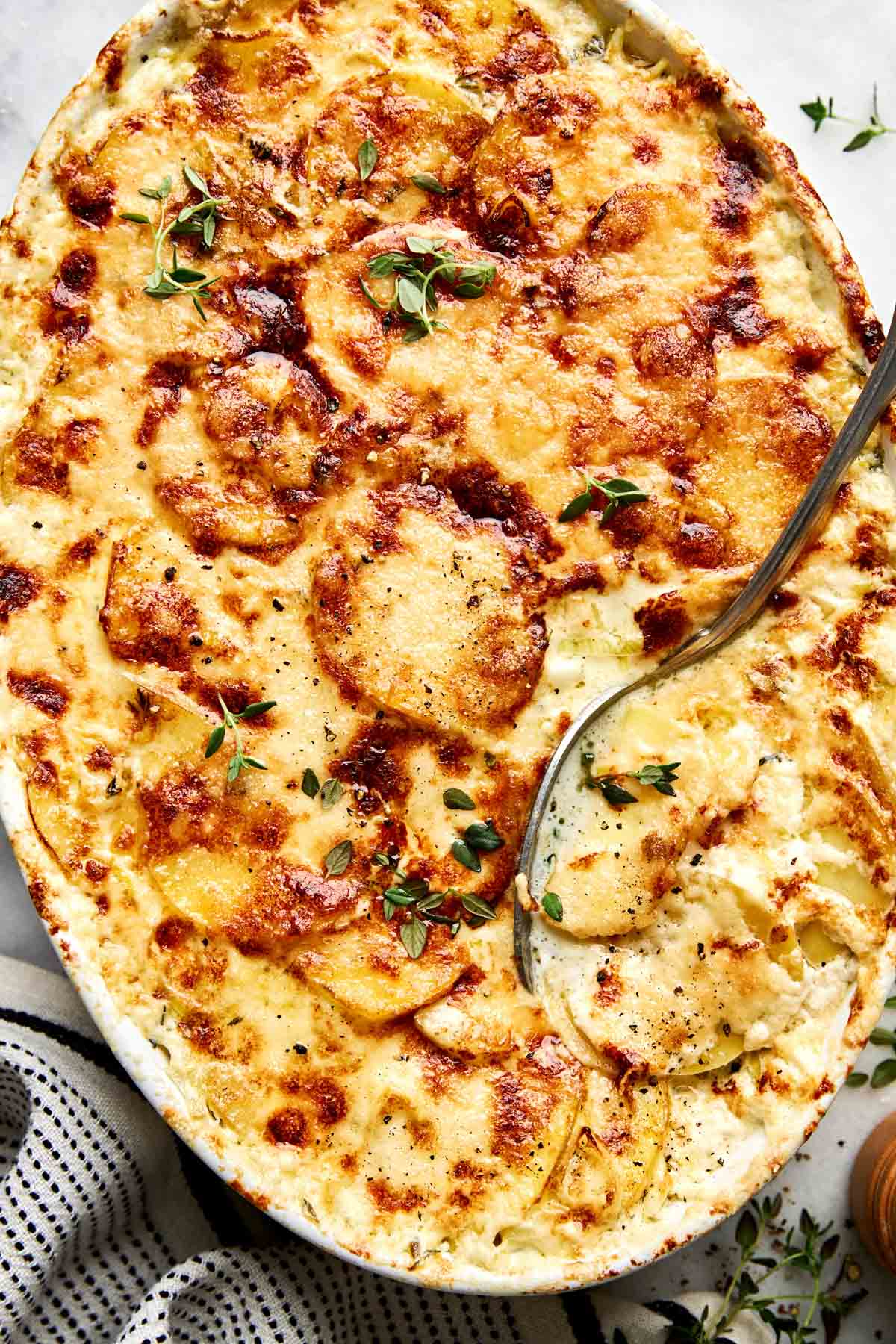 A baking dish of au gratin potatoes with a browned cheese crust, topped with fresh herbs and a serving spoon.
